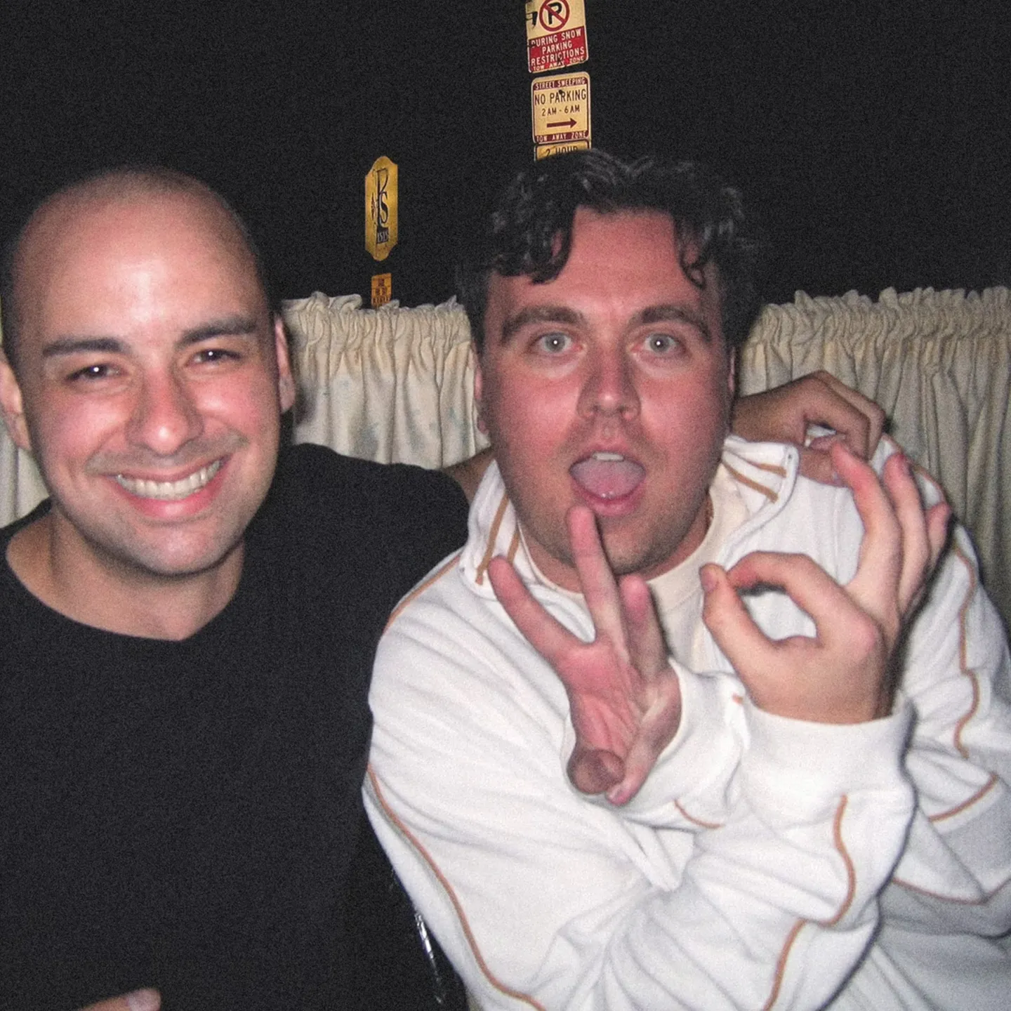 Two men pose playfully in a dimly lit room with curtains in the background. The man on the left smiles broadly, while the man on the right makes a hand gesture.