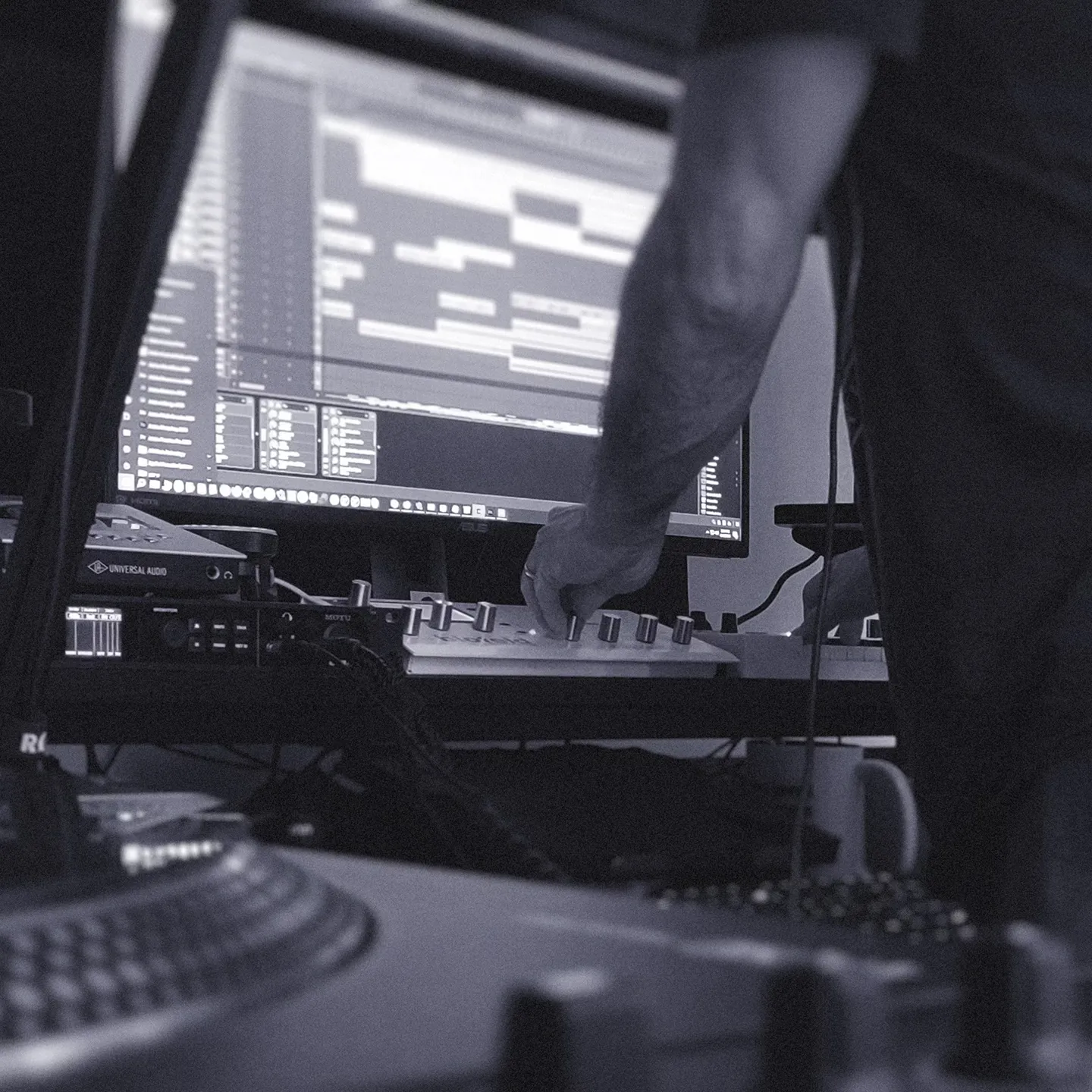 A person using music production equipment in a studio setting, with computer screens displaying software and audio tracks in the background.
