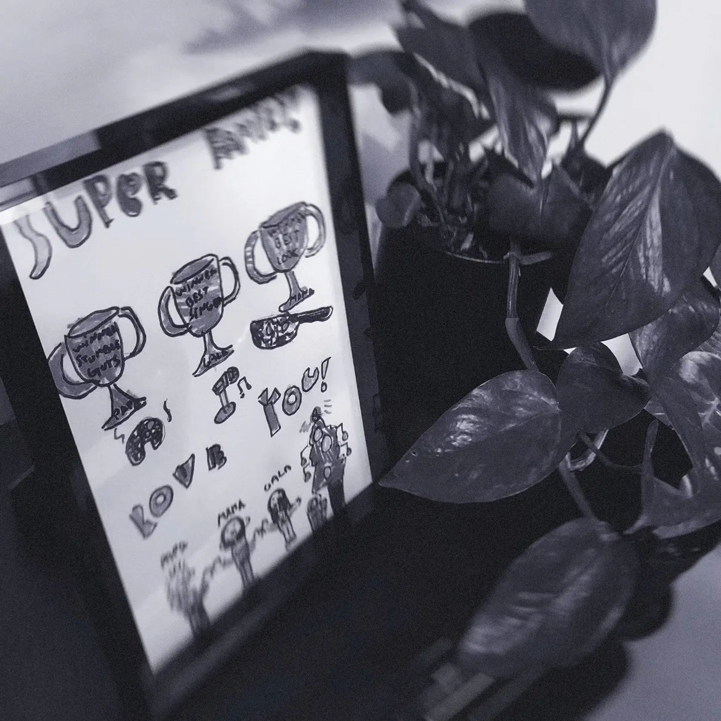 Framed child’s drawing of trophies with text "Super Family" and "I Love You!" next to a leafy plant, conveying encouragement and love.