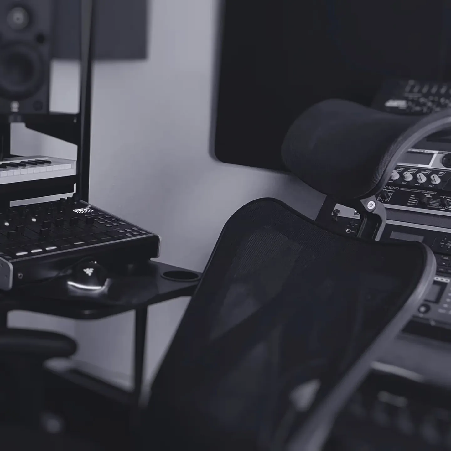 A black mesh office chair is positioned in front of audio equipment, including a mixer, speakers, and rack-mounted gear in a music studio.